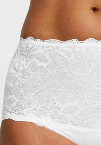 Close-up van de taille van een persoon die witte kantonderbroek draagt met bloemmotieven en een golvende rand op een neutrale achtergrond.