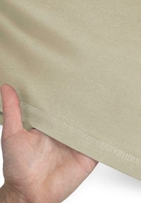 Hellbeige Baumwollstoff, der von einer Hand gehalten wird. Der Stoff zeigt eine glatte Textur und sichtbare Nähte entlang der Kante.