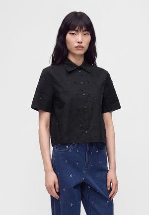 Femme portant une chemise noire à manches courtes boutonnée avec des détails en perles et un jean bleu avec de petits motifs blancs, debout sur un fond uni.