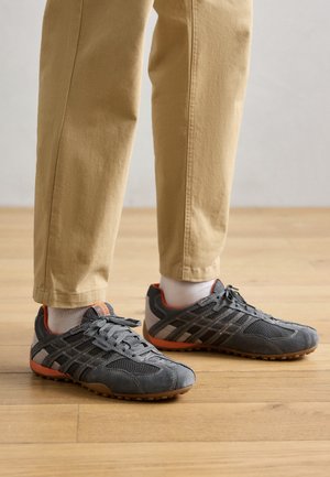 Szürke, könnyű sportcipők, hálós és velúr anyagokkal, narancssárga részletekkel a talpon és a sarkon, valamint szürke fűzőkkel, világos bézs nadrággal párosítva.