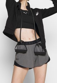 Černá mikina na zip se stříbrným logem, černá sportovní podprsenka a šedé šortky s černým pasem. Modelka drží fitness odporové gumy.