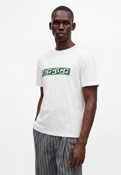Jeune homme portant un t-shirt blanc à manches courtes avec un texte graphique vert et noir et un pantalon gris rayé, debout sur un fond blanc.