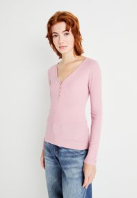 Langermet, ribbet rosa topp med V-hals og tre knapper foran. Akkompagnert av blå denimjeans.