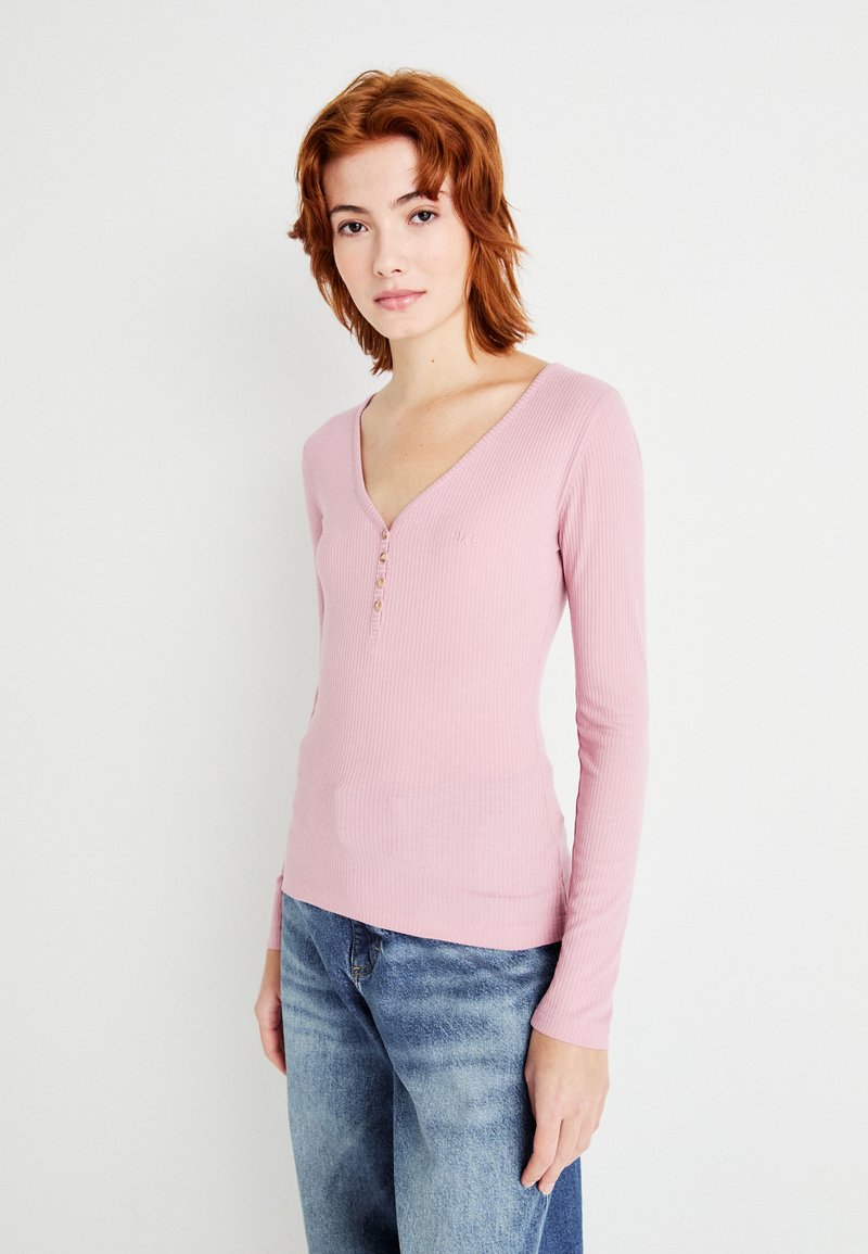 Langermet, ribbet rosa topp med V-hals og tre knapper foran. Akkompagnert av blå denimjeans.