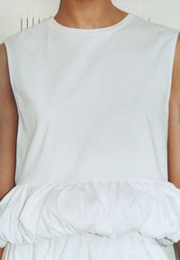 Femme portant une robe blanche sans manches avec un détail de taille à volant de style peplum sur un fond neutre.