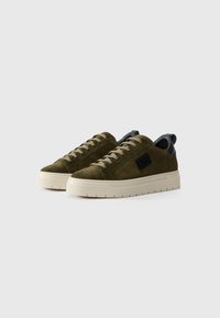 Ein Paar olivgrüne Wildledersneaker mit dicken beigen Sohlen und Schnürsenkeln, mit einem schwarzen ledernen Fersenbereich, die nebeneinander auf einem weißen Hintergrund platziert sind.