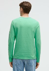 Langärmliges grünes Shirt mit einer strukturierten Oberfläche und Rundhalsausschnitt. Verfügt über einen schwarzen Grafikdruck am linken Ärmel. Kombiniert mit hellblauen Jeans.