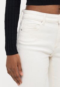 White denim jeans with a fitted shape, featuring a smooth texture, front pockets, and a top button closure. Black ribbed long-sleeve shirt visible.
