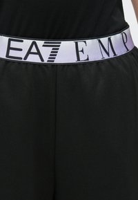 Čierne športové šortky majú biely elastický pás so zvýrazneným nápisom "EA7 EMP" a gradientným vzorom, čo zdôrazňuje textúru a dizajn.