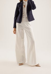 Blazer azul marino con solapas de muesca, blusa blanca con corbata y pantalones blancos de pierna ancha. El conjunto se completa con zapatos planos beige con lazos.