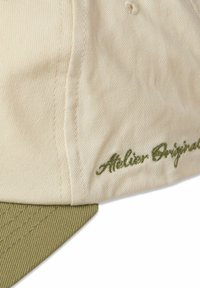Beige cap with a textured fabric, green underside on the visor, and green embroidery reading "Atelier Original."
