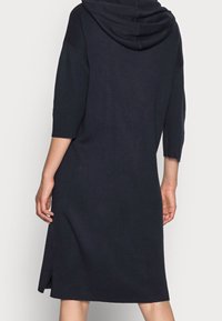 Mörk marinblå klänning i stickad tröja med huva, trekvarts ärmar, sidoflikar och ett mjukt, texturerat tyg. Enkel, rak silhuett.