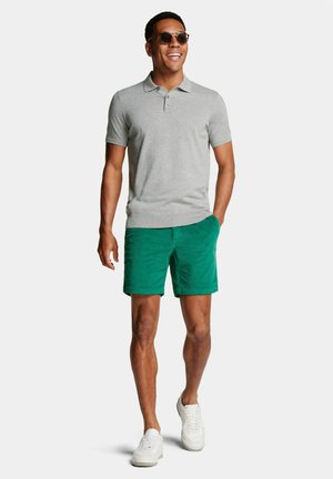Homme portant des lunettes de soleil, un polo gris à manches courtes, un short vert et des baskets blanches, marchant avec une main dans la poche et souriant.