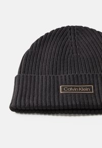 Czarna dzianinowa czapka beanie o strukturalnym splocie. Posiada podwinięty brzeg i prostokątną metkę z logo Calvin Klein w złotym kolorze z przodu.