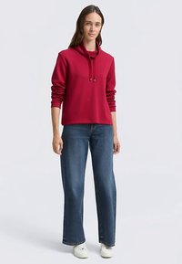 Red hoodie with a soft texture, drawstring neckline, and ribbed cuffs, paired with loose-fitting blue jeans and white sneakers.