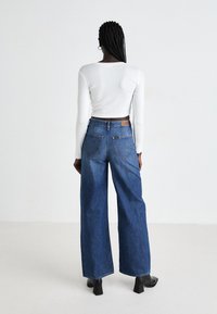 Una donna con capelli intrecciati è in piedi di fronte a un muro bianco, indossando un top corto bianco a maniche lunghe, jeans blu a gamba larga e stivali neri con tacco.
