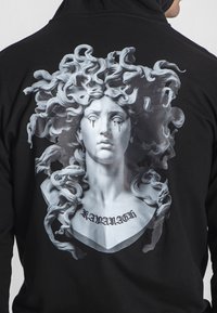 Crna dukserica s velikim, printom u nijansama sive boje koji prikazuje klasični kip s valovitom kosom i naglascima u obliku suza, s natpisom "KAVANAGH" ispod.