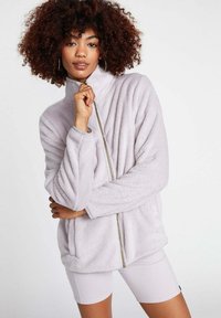 Giacca zip-up di un morbido viola chiaro, con colletto alto e tessuto texture. Presenta maniche lunghe e un dettaglio della zip dorata in contrasto.