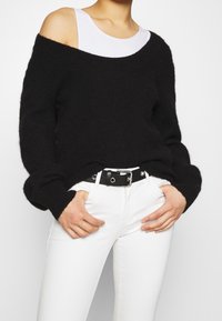 Suéter negro sin mangas hecho de material suave, combinado con jeans blancos. Presenta puños acanalados y un cinturón negro con detalles en hardware plateado.