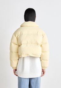 Korte gele pufferjack met een hoge kraag en brede, gewatteerde structuur. Draagend over een witte geribbelde kledingstuk heeft het een cropped ontwerp.