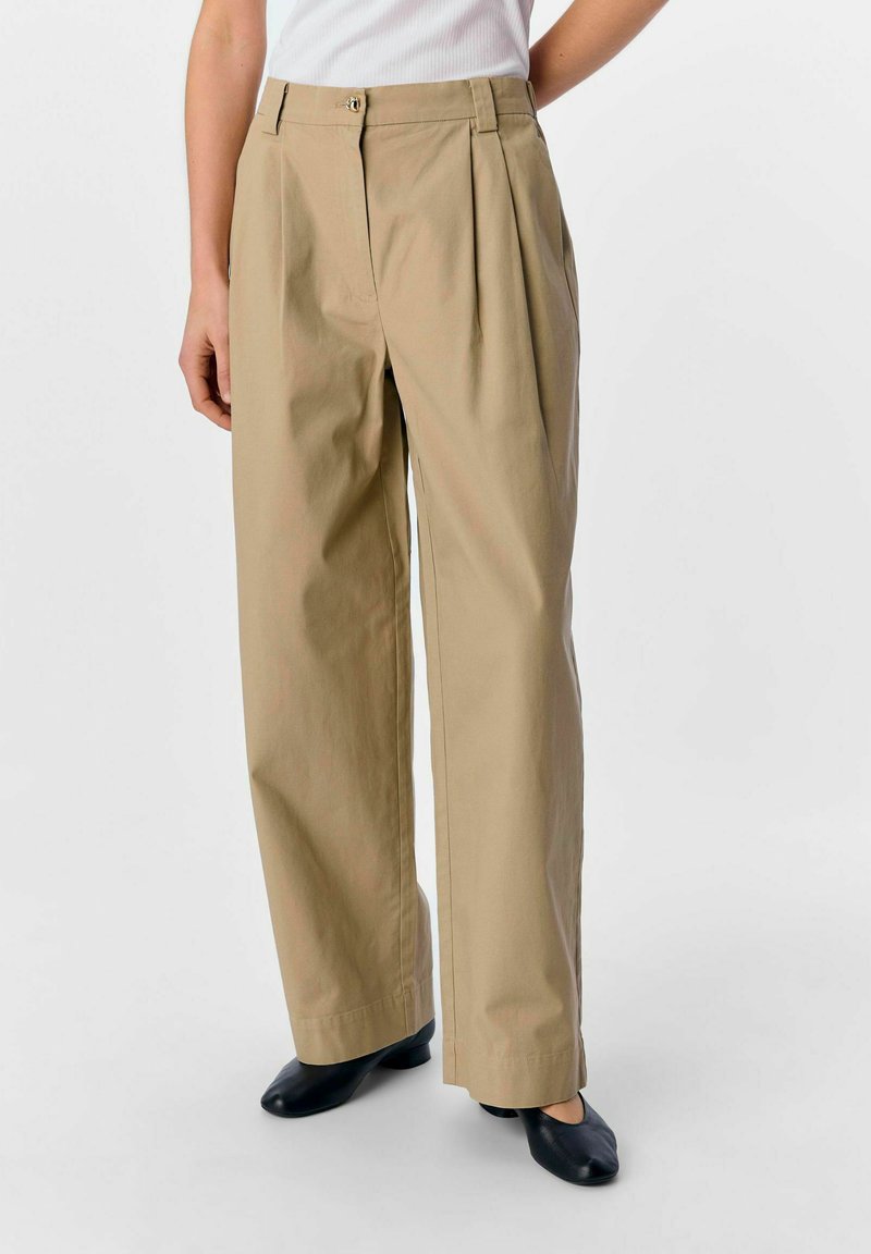Pantalons larges beiges avec plis avant, fabriqués en tissu de mélange de coton. Ils sont dotés d'une fermeture éclair et d'un bouton, associés à des chaussures noires.