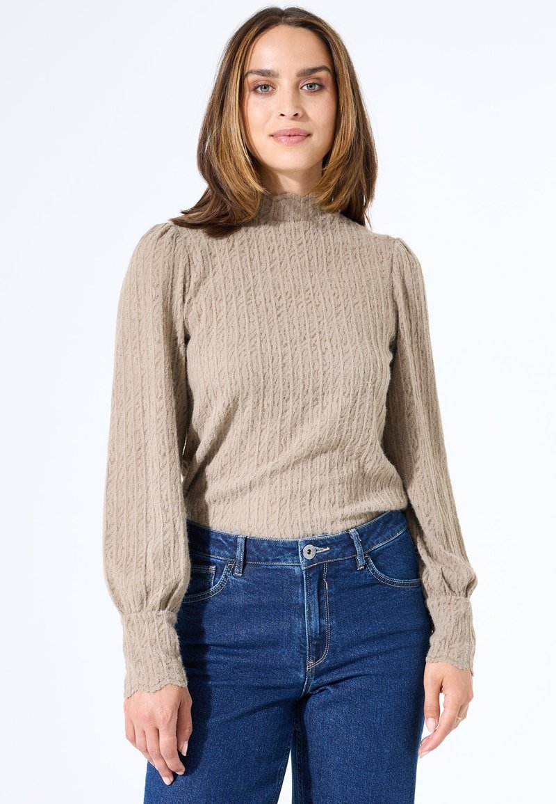 Beige strukturiertes langärmliges Oberteil mit hohem Kragen und puffigen Schultern, kombiniert mit dunkelblauen Jeans mit vorderem Knopfverschluss.