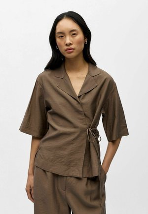 Žena s dlhými tmavými vlasmi má na sebe hnedý zavinovací top s krátkym rukávom a golierom a k nemu ladúce voľné nohavice, stojaca pred bielym pozadím.