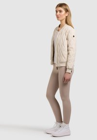 Jeune femme portant une veste matelassée beige, une chemise blanche, des leggings beiges et des baskets montantes blanches, debout devant un fond uni.