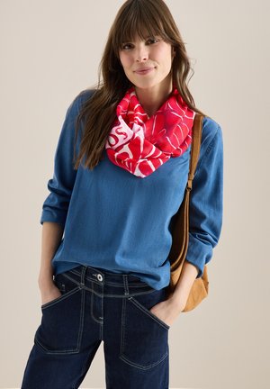 Vrouw draagt een blauw shirt met lange mouwen, donker denim broek, een rode sjaal met patroon en een bruine schoudertas, staand met handen in de zakken.