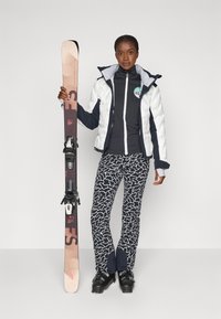Equipamiento de esquí que incluye una chaqueta acolchada blanca y azul marino, una sudadera azul marino con cremallera, pantalones a rayas en blanco y negro, botas de esquí negras y esquís con fijaciones.