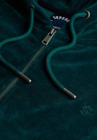 Sudadera verde de tela suave con cremallera, superficie texturizada, cordones ajustables en la capucha y una etiqueta con el logo. Presenta una cremallera metálica.