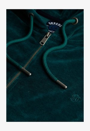 Green, soft fabric hoodie with a zipper, textured surface, adjustable hood cords, and a logo tag. Features a metallic zipper.