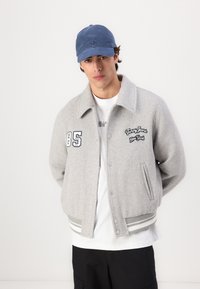 Giubbotto bomber grigio in lana con polsini a righe, numeri e logo ricamati. Sotto c'è una maglietta bianca e un cappellino blu completa il look.