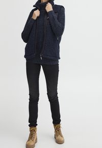 Osoba oblečená v navy modré texturované vestě a svetru, černých slim kalhotách a světlých šněrovacích botách stojí před jednoduchým pozadím.