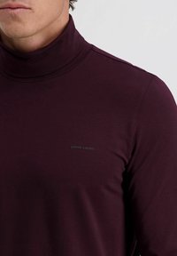 Mann trägt einen eng anliegenden, dunkelburgundfarbenen Rollkragenpullover mit einem kleinen "Pierre Cardin"-Logo auf der linken Brust.