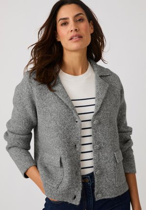 Vrouw met een grijze wollen jas over een wit shirt met zwarte horizontale strepen en donkerblauwe spijkerbroek, poseert met één hand in haar zak.