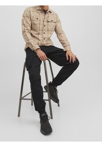 Beige kostkovaná košile se dvěma náprsními kapsami, spárovaná s černými cargo kalhotami. Model sedí na kovové stoličce a má na sobě černé tenisky.