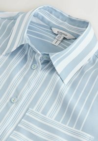 Chemise bleu clair à col avec des rayures verticales blanches, des boutons bleus et une poche poitrine avant, présentée à plat sur un fond pâle.