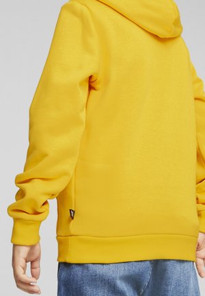Sweat à capuche jaune en tissu texturé, avec une grande capuche, des poignets côtelés et un ourlet. Petite étiquette de marque visible sur le côté inférieur.