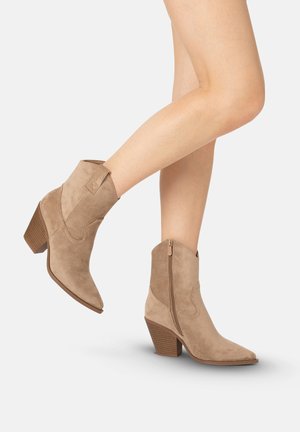 Ben iført lysebrune ruskind ankelstøvler med stablet blokhæl og sidelås, placeret mod en ensfarvet lys baggrund.