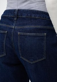 Κοντινό πλάνο σκούρου μπλε τζιν με ορατές πίσω τσέπες και κίτρινη ραφή, φορεμένα με ανοιχτό γκρι πουκάμισο.