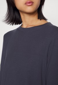 Chemise gris foncé à manches longues avec un col rond. Texture douce ; le tissu semble léger et légèrement extensible. Pas de motifs ni d'accents visibles.