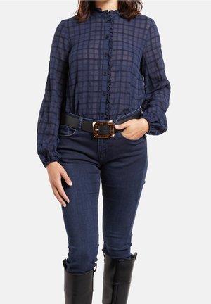 Femme portant un chemisier à carreaux bleu marine avec une patte de boutonnage à volants, un jean foncé, une ceinture noire avec une grande boucle et des bottes noires montantes jusqu'aux genoux.