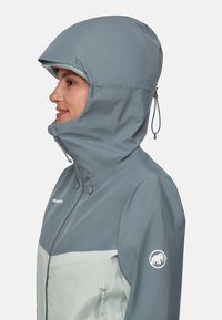 Kvinne iført en to-farget grå vanntett jakke med hette, glidelås foran og logo på ermet, vendt mot venstre i profil.