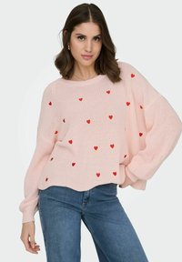 Femme portant un pull en maille rose clair avec des motifs de cœurs rouges et un jean bleu, posant avec la main sur la hanche, fond neutre.