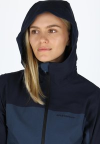 Navy blue waterproof jacket with a hood, featuring a lighter blue panel at the chest, front zipper, and logo on the left side.