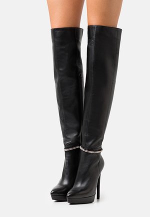 Botas de tacón stiletto de cuero negro, hasta la rodilla, con suelas de plataforma y detalle de cadena plateada alrededor de los tobillos, usadas sobre piernas desnudas.