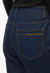 Mørk denimjeans med oransje sømmer og en baklomme med brodert tekst "La dame de cœur." Skreddersydd passform og subtil tekstur.