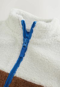 Pile con mezzo zip in colore panna e marrone, materiale testurizzato, caratterizzato da una zip blu e un design a blocchi di colore a contrasto.