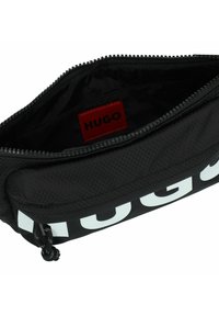 Zwarte stoffen tas met een gestructureerde afwerking, voorzien van een opvallend wit "HUGO" logo, een ritsluiting en een rode binnenlabel.